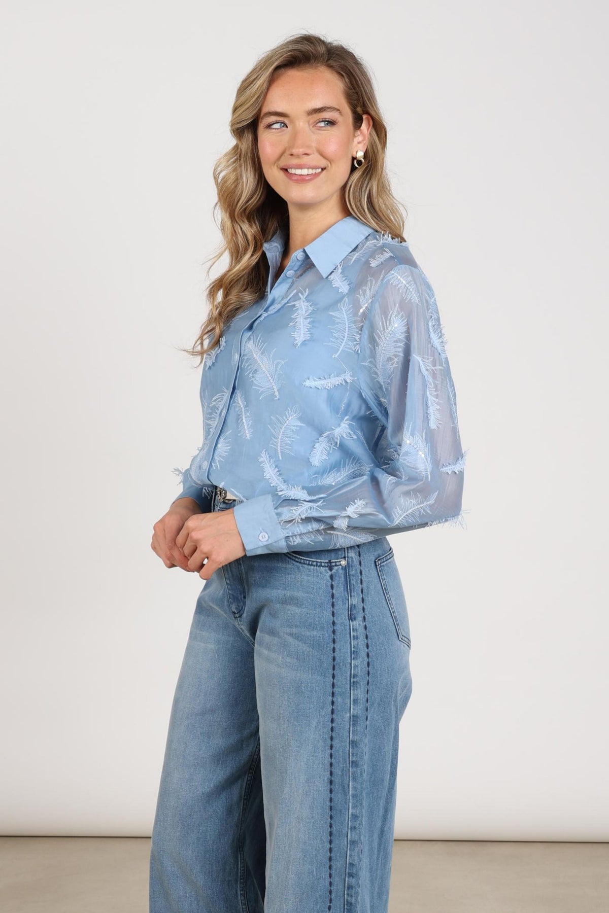 Lichte blauwe blouse Aysha met verenpatroon van Nukus, casual en stijlvol, hangend aan een houten haak.